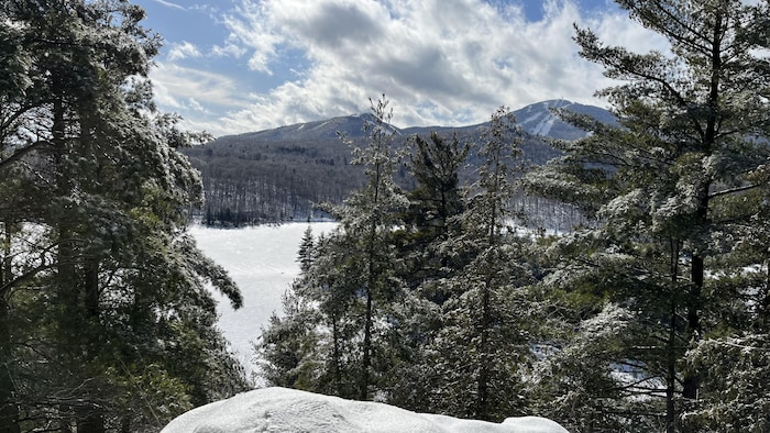 Derrière des sapins, on voit lac gelé au pied d'une montagne sillonnée par des pistes de ski.