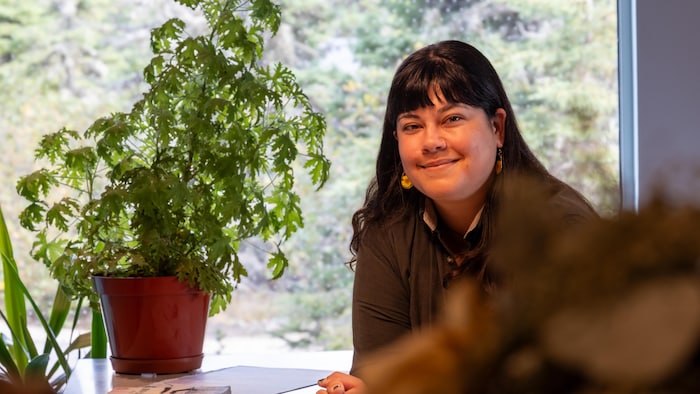 Une femme accoudée à une table sourit près d'une plante verte.