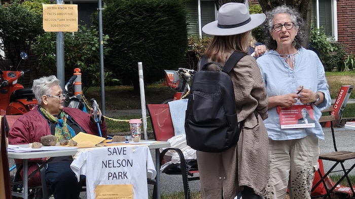 Des gens se parlent dans un parc, devant une table où une pancarte dit « Sauvez le parc Nelson ».