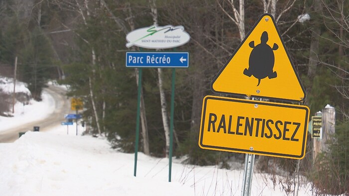 Une pancarte jaune où l'on peut voir une image de tortue et le mot «ralentissez» devant un écriteau annonçant l'entrée du Parc récréotouristique. 