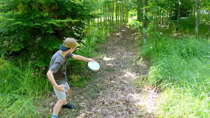 Un jeune portant une casquette lance un frisbee dans un sentier en forêt.