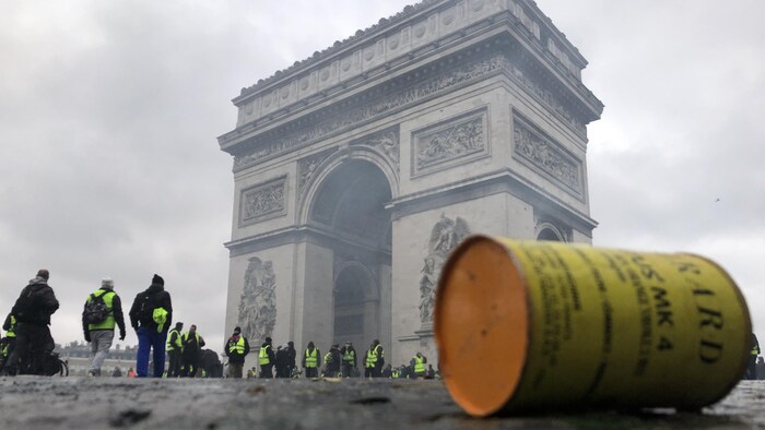 Une canette de gaz lacrymogène au sol est visible à l'avant-plan devant des manifestants portant des gilets jaunes debout devant l'Arc de triomphe, à Paris.