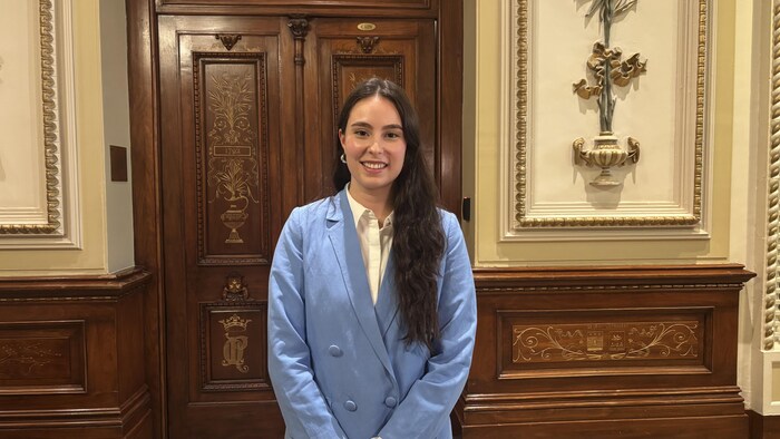 Une jeune femme souriante, aux longs cheveux bruns, pose debout devant une porte en bois sculpté. Elle porte une chemise blanche sous une veste de tailleur bleu clair. Le décor est richement orné de moulures et de dorures classiques.