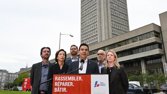 Charles Milliard s'exprime à un lutrin du PLQ, entouré de cinq candidats, devant une grande tour en béton.