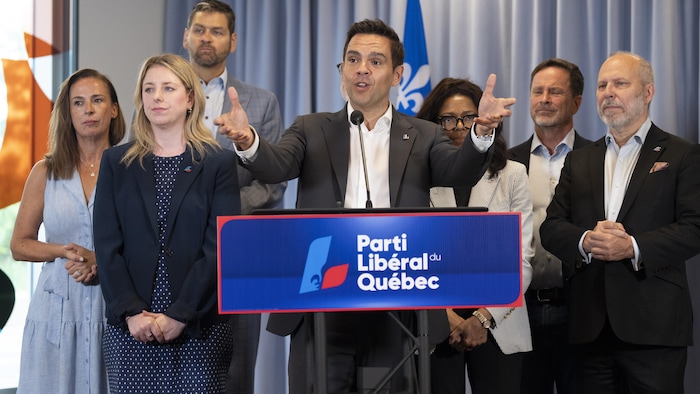 Charles Milliard parle au micro derrière un pupitre du Parti libéral du Québec, entouré de personnes.