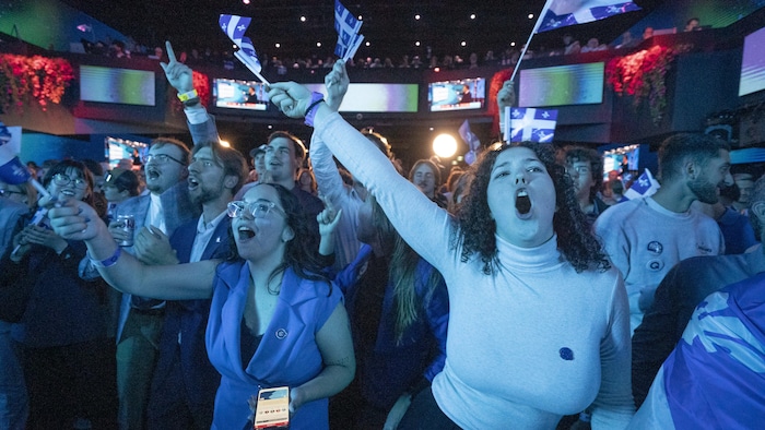 Une foule en liesse brandit des drapeaux du Québec dans une salle intérieure.