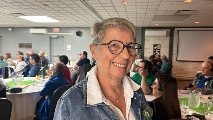 Une femme souriante. Derrière elle, des gens assis autour de tables participent à l'Assemblée générale annuelle 2025 du Parti Vert du Nouveau-Brunswick.