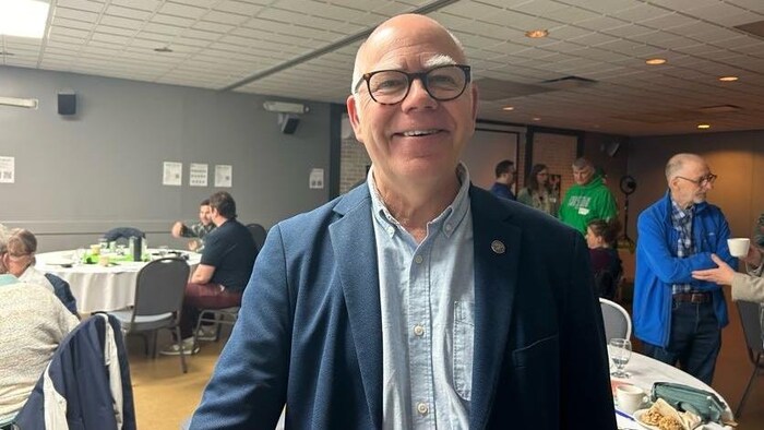 Un homme souriant. Derrière lui, des gens assis autour de tables participent à l'Assemblée générale annuelle 2025 du Parti Vert du Nouveau-Brunswick.