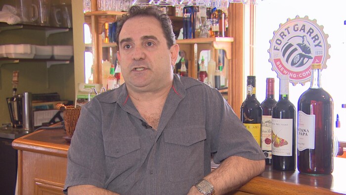 Joe Loschiavo accoudé à un bar en bois sur lequel se trouvent des bouteilles d'alcool.