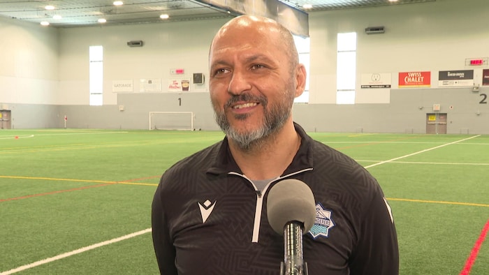 Patrice Gheisar, souriant, les mains dans le dos avec un micro sur pied devant lui, est debout sur les lignes de côté d'un terrain de sport intérieur.