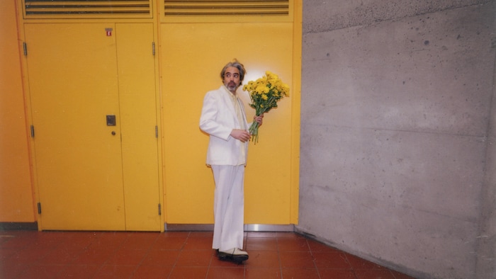 Un homme portant un costume blanc tient un bouquet de fleurs jaunes. 