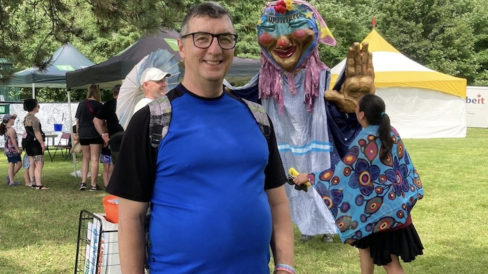 Paul-André Gauthier pose pour une photo sur le site du festival tandis qu'une marionnette géante se tient derrière lui.