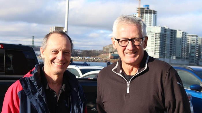 Paul Hines et Geoff Lebans sourient à la caméra dans un stationnement avoisinant un port.