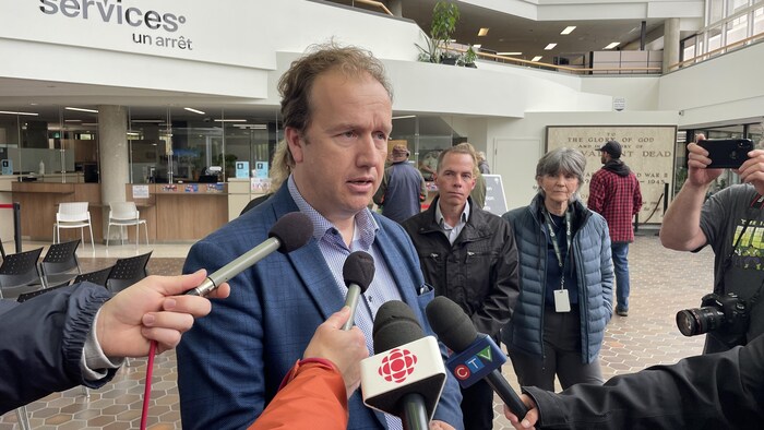 Paul lefevbre devant plusieurs microphones avec trois personnes derrière lui.