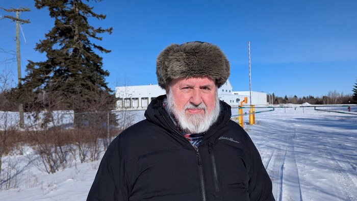 Debout dans un paysage hivernal, un bâtiment industriel derrière lui, un homme à la barbe blanche est coiffé d'un impressionnant chapeau de poil.