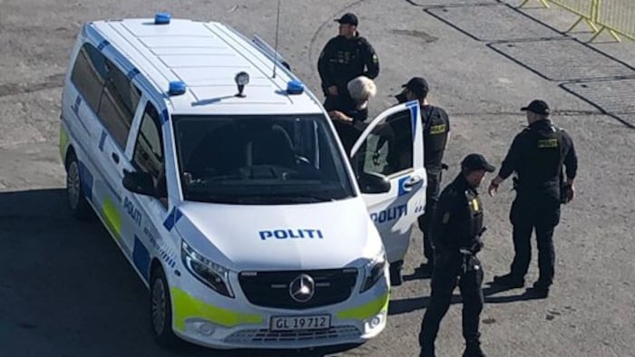 A man is placed on the side of a police car. Police officers are facing him, while others nearby are watching the surroundings.