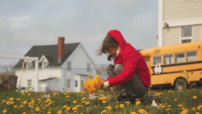 Une jeune fille accroupie cueille des pissenlits devant une maison et un autobus scolaire sur l'île d'Anticosti.