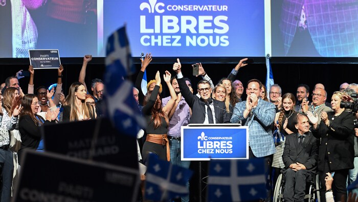 Éric Duhaime lève son pouce sur la scène du hall du Centre Vidéotron, entouré de ses candidats et devant une foule. 