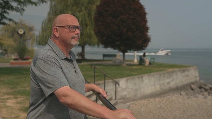 Un homme chauve portant des lunettes et un polo gris est appuyé sur une rampe, de profil, le regard tourné vers la gauche. Derrière lui, une promenade en bord de lac.