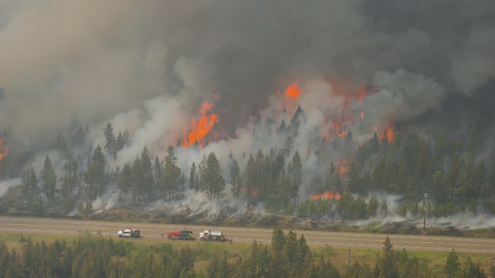 Vue aérienne d'une forêt enfumée et en flammes, le long d'une route à plusieurs voies, où se trouvent des camions spécialisés.