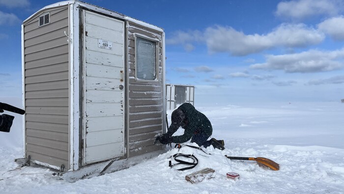 Une personne est agenouillée près d'une cabane à pêche sur une étendue couverte de neige.