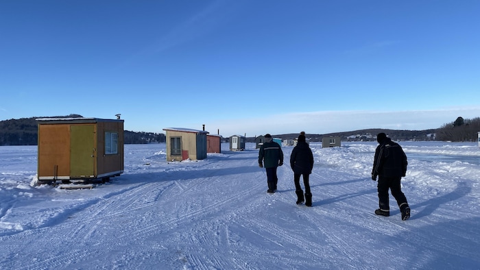 Trois personnes marchent sur une route déneigée sur un lac. De part et d'autre de la route, de petites cabanes de pêche colorées.