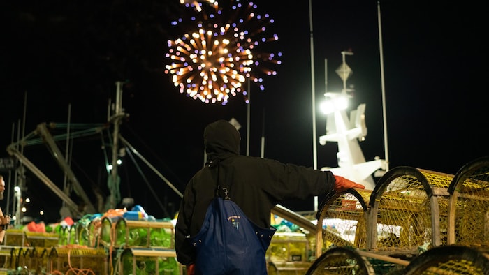 Un pêcheur regarde un feu d'artifice. Il a la main posé sur un empilement de casiers à homards.