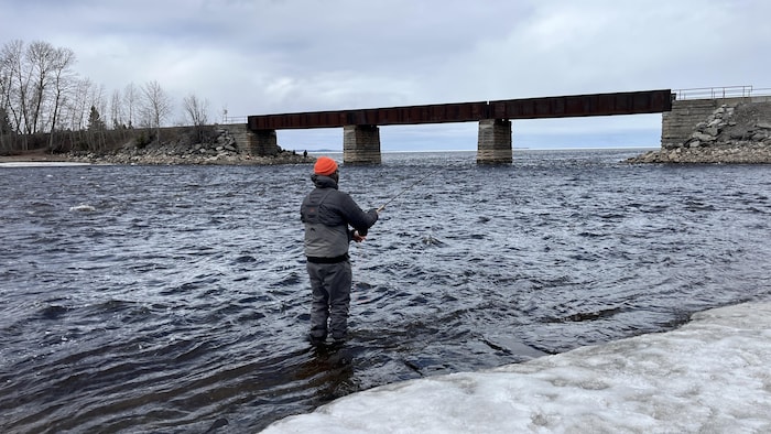 Un homme de dos pêche dans une rivière. Les arbres sur l'autre rive sont dénudés et de la glace est visible sur le bord.