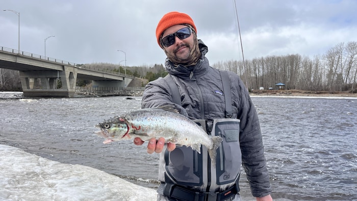 Un homme souriant portant des vêtements chaud tient à une main un poisson. Une rivière et un pont sont visibles derrières lui. 