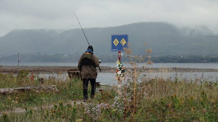 Un pêcheur amateur marche vers la pointe du barachois, où une affiche transformée en mémorial indique les dangers du secteur.