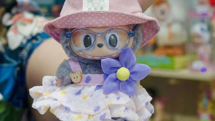 Une main   tendu montre une porte-clef Labubu accessoirisée d'une salopette, de chaussure de people   et d'un chapeau.