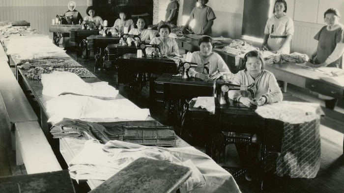 De jeunes filles sont réunies dans une salle de couture assise derrière leurs machines à coudre et entourées de tissus et de vêtements disposés sur de longues tables de travail.