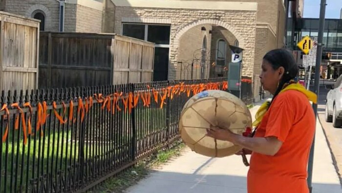 Gerry Shingoose toca el tambor y canta frente a la valla de la catedral de Santa María, donde cuelgan cintas naranjas.