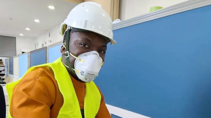 Un homme accroupi portant un casque de sécurité blanc, un masque de protection N95 et un gilet jaune haute visibilité trempe un pinceau dans un pot de peinture blanche. Il regarde vers l'objectif tout en travaillant devant un mur bleu orné de moulures blanches.