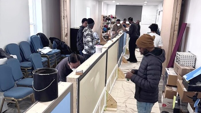 Plusieurs personnes s'affairent autour de cloisons sèches temporaires installées au centre d'une pièce étroite. Les participants, certains portant des masques de protection et des vêtements d'hiver, travaillent sur les finitions des murs, y compris la peinture bleue et la pose de moulures blanches.