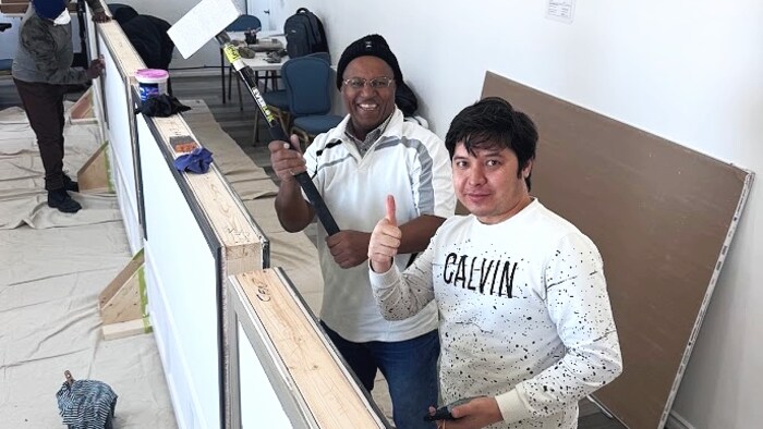 Darren WeasleChild et un de ses étudiants qui tient une ponceuse de gypse à long manche sourient devant une structure de cloisons sèches en construction dans un atelier de formation.