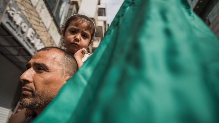 Une petite fille est juchée sur les épaules de son père derrière un drapeau du Hamas.