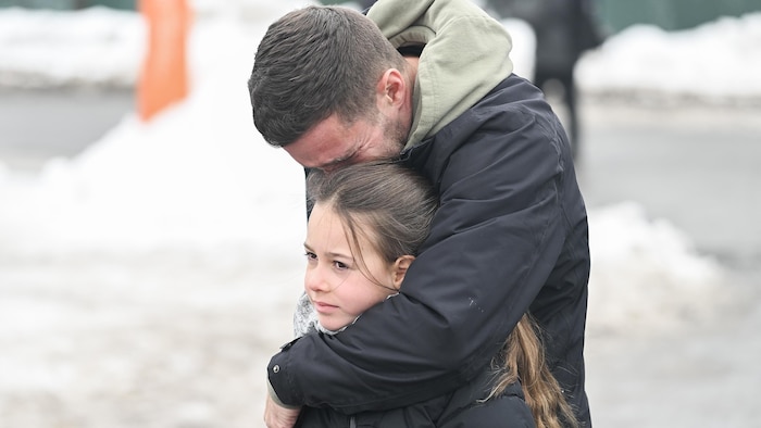 Un père en pleurs embrasse sa fille en la serrant dans ses bras, pendant que l'enfant regarde au loin, l'air triste.