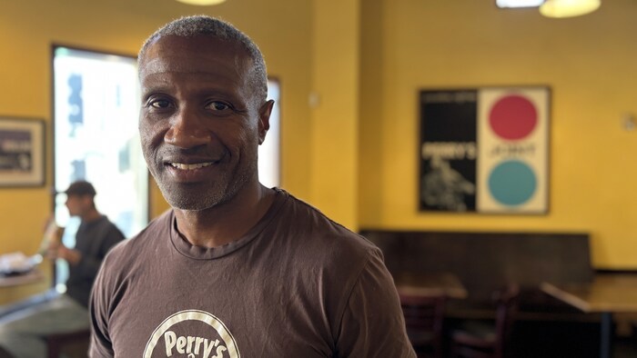 Dans la salle à manger de son restaurant, Perry Bennet, un homme noir qui porte un t-shirt à l'effigie de son entreprise, regarde la caméra. 