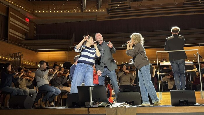 Trois personnes chantent sur scène devant un orchestre. 