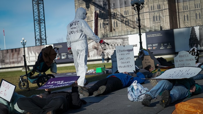 Une personne fait semblant de disperser un pesticide sur des personnes au sol faisant semblant d'être mortes.