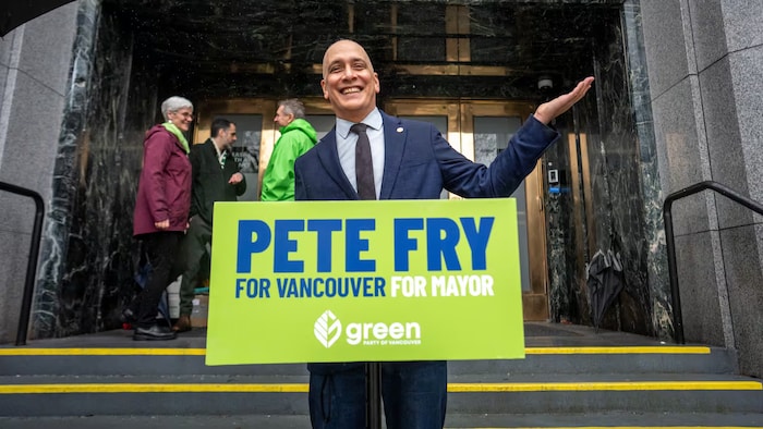 Ken Fry derrière un pupitre lève le bras gauche.