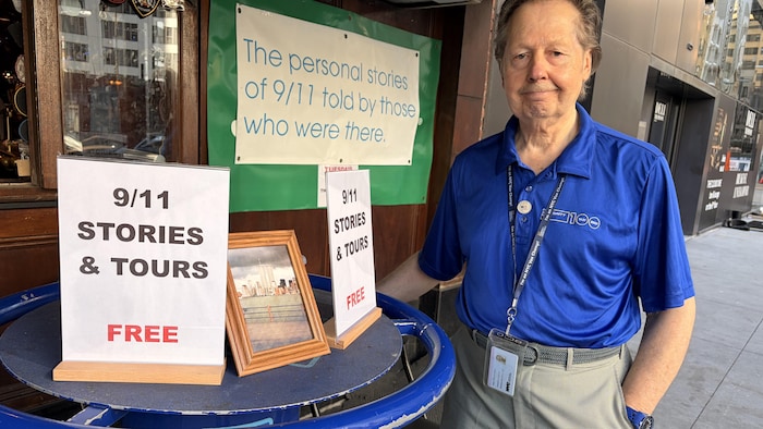 Un homme debout devant un stand sur lequel il est installé des pancartes et une photo des anciennes tours jumelles du World Trade Center.