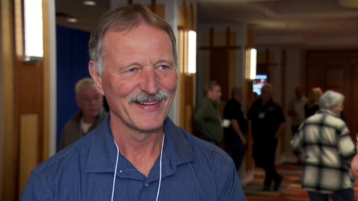 Un homme à la moustache soigneusement taillée et aux yeux bleus et ronds comme des bleuets esquisse un large sourire.