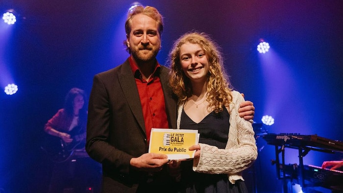 Une jeune fille souriante sur scène accompagnée d'un adulte pose avec un carton sur lequel est écrit Prix du public du petit gala de la chanson de Caraquet. 