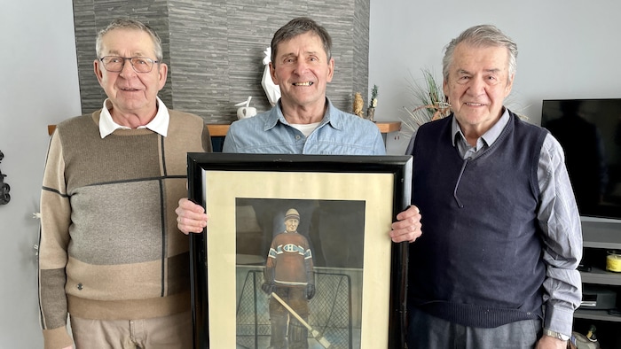 Trois hommes prennent la pose dans une pièce et l'homme au centre tient un cadre sur lequel est représenté un gardien de but de hockey. 