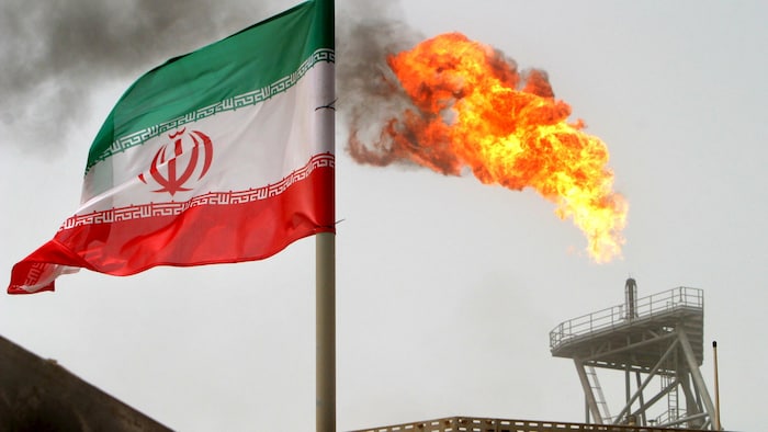 The Iranian flag floats in the wind near a torch of an oil well from which a column of fire escapes. falling price gasoline quebec