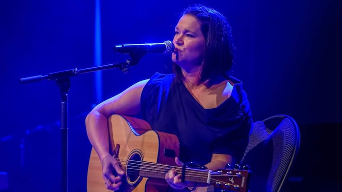 Une femme assise chante et joue de la guitare devant un micro.