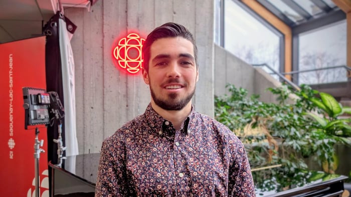 Un homme vêtue d'une chemise fleurie prend la pose devant un logo de Radio-Canada. 