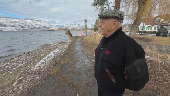 Colin Pritchard est à l'extérieur, devant un lac et des montagnes. Il y a un peu de neige sur le sol. 